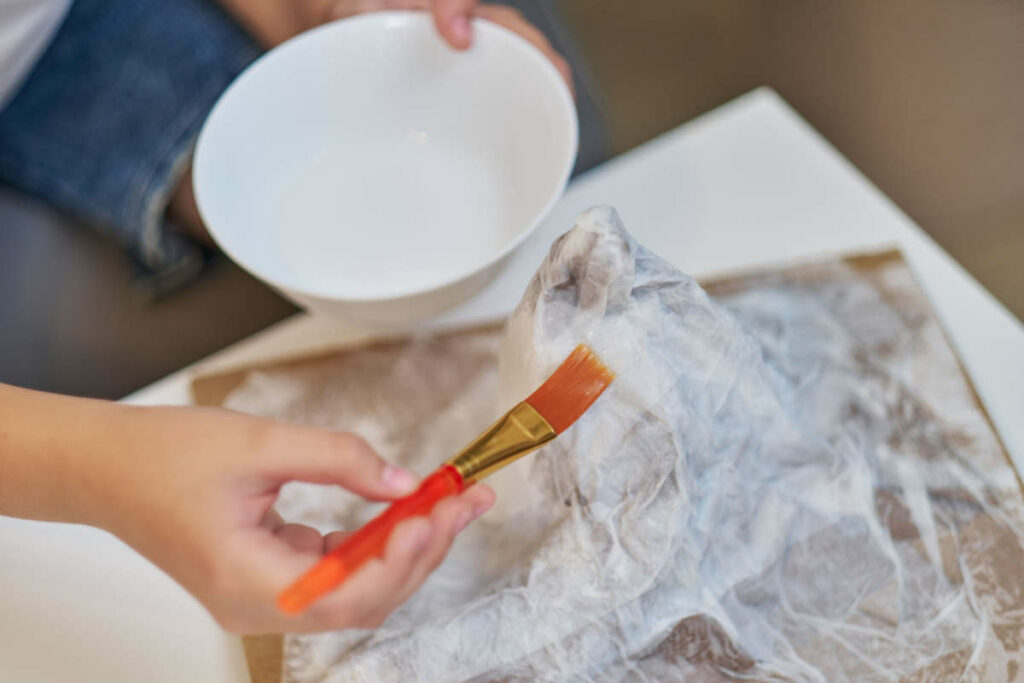 mani di bambino con una bacinella d'acqua, un pennello e dei fogli di giornale che crea la cartapesta