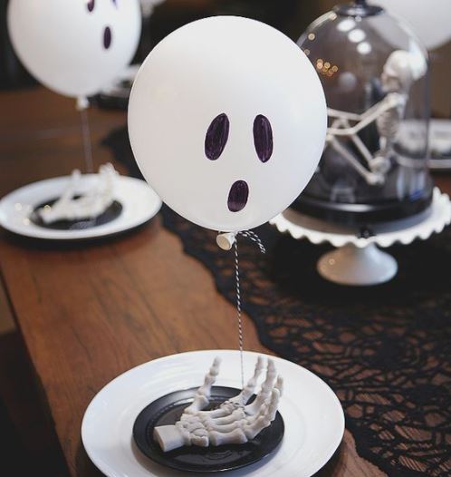 tavola allestita per halloween con palloncini bianchi con occhi e bocca disegnata, su un piatto nero con una mano finta poggiata