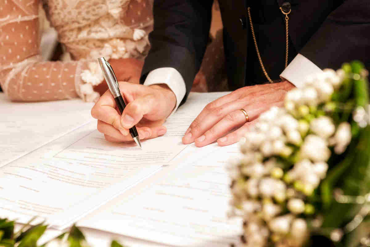 mani di uomo sposato che firmano gli atti matrimoniali, in secondo piano braccia della sposa con abito