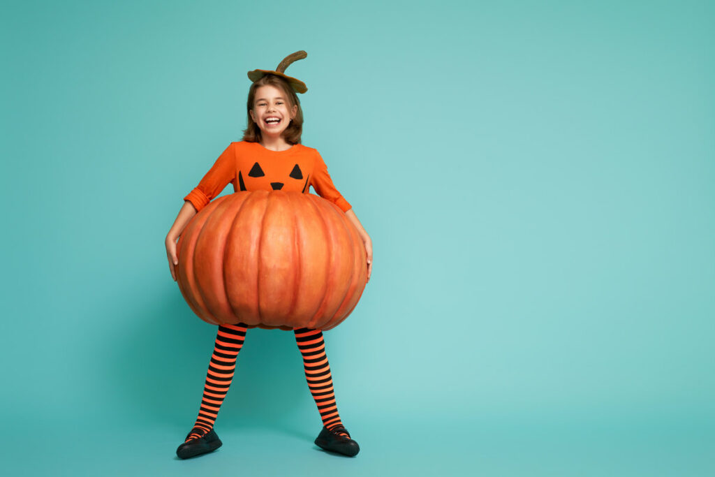 bambina mascherata da zucca, con calze a righe di colore nero e arancio, maglia arancione e zucca come gonna su sfondo azzurro