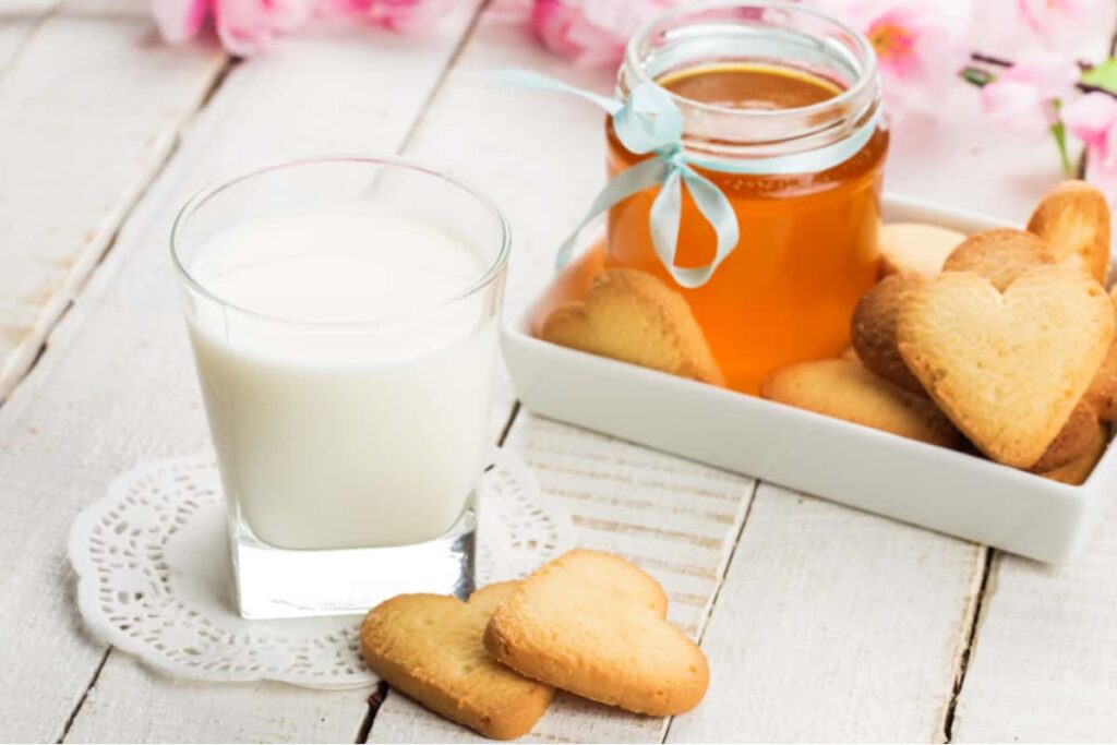bicchiere di latte bianco con dei biscotti a forma di cuore in un vassoio con il miele