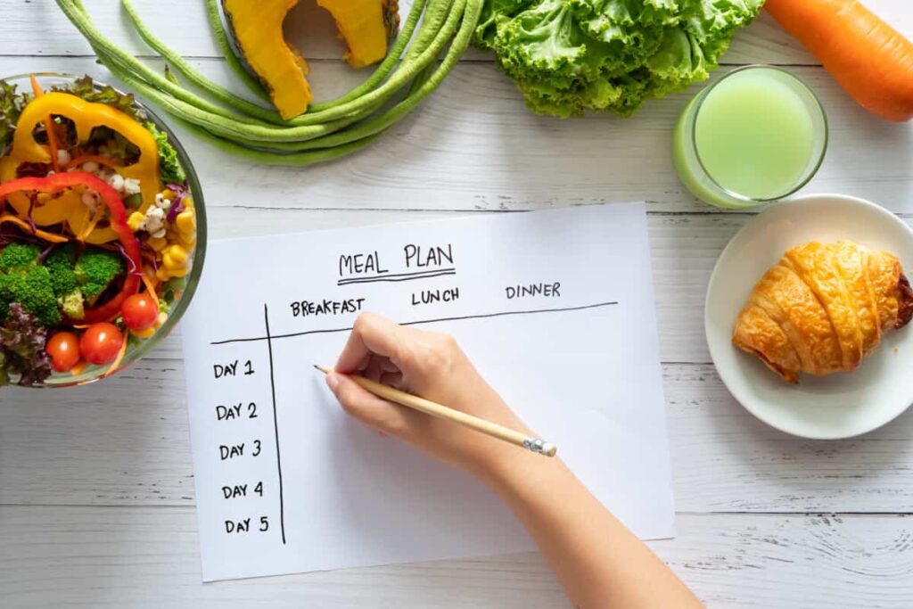 mani di ragazza che scrivono una tabella con i giorni della settimana per il piano alimentare, su un tavolo, con frutta e verdura intorno