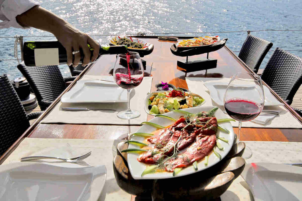 tavola apparecchiata con cibo al centro della tavola, due bicchieri con mano di ragazza che versa vino rosso, con il mare sullo sfondo