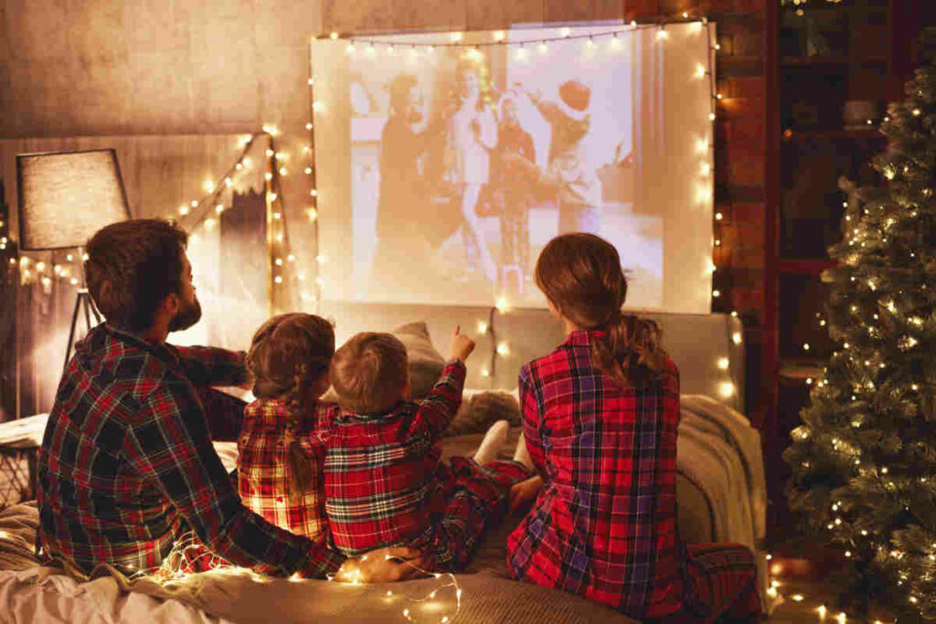 famiglia composta da mamma, papà, bambino, bambina tutti con il pigiama a quadri rosso e verde, seduti sul divano mentre guardano un film di natale, circondati da atmosfera natalizia con lucine