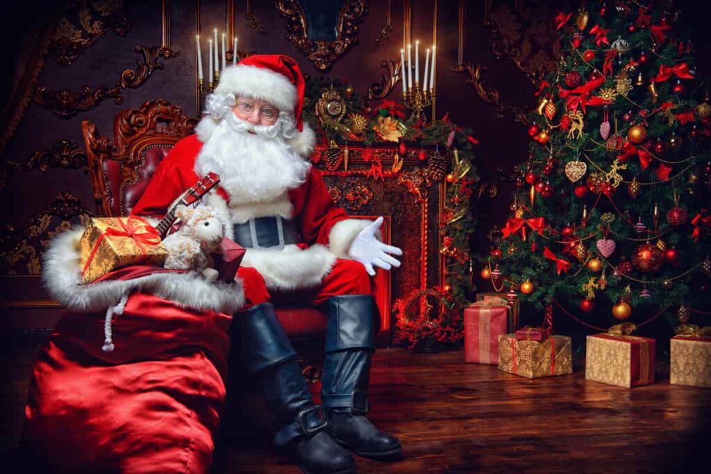 babbo natale vestito di rosso, con barba bianca, seduto, con regali intorno e albero di natale sullo sfondo