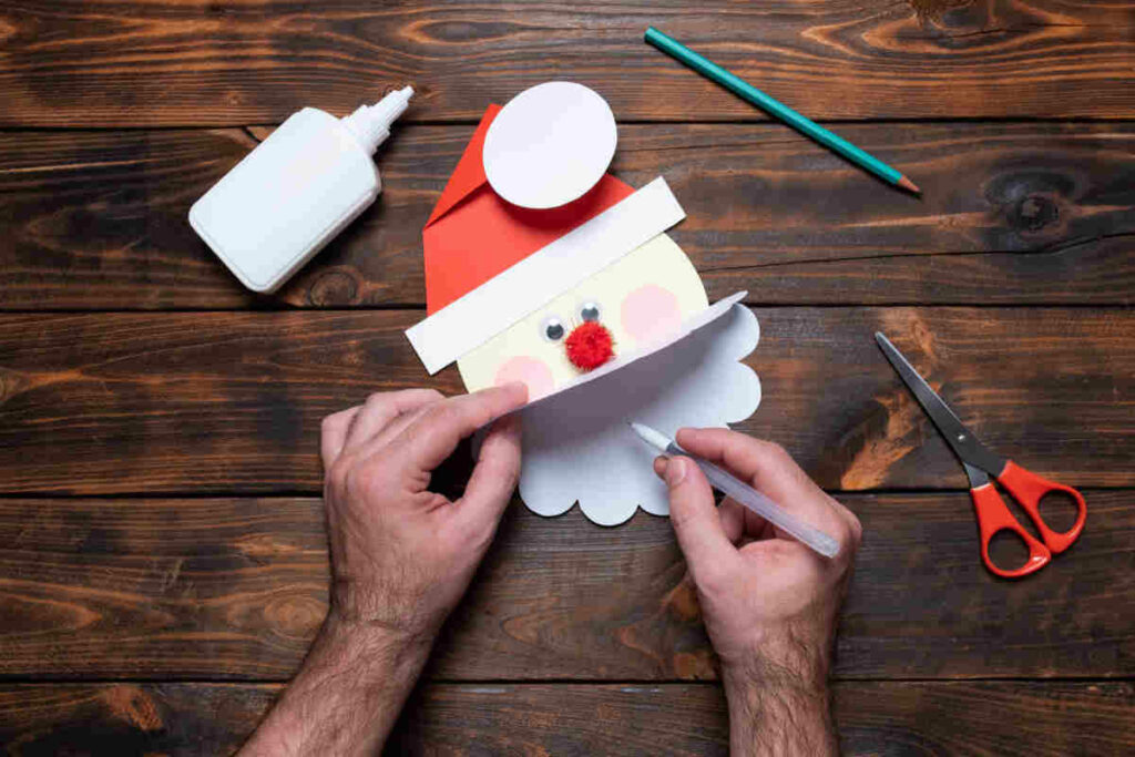 mani di uomo che scrivono un bigliettino di natale con la forma del volto di babbo natale, poggiato su un tavolo in legno con oggetti come forbici e colla
