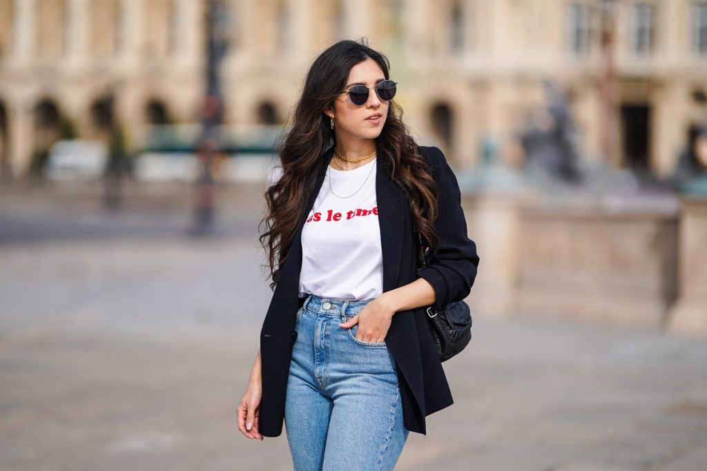 Camille Grandguillote wears sunglasses, a black blazer jacket from Sezane, a black leather bag from Kate Lee, a white t-shirt from "Pas le Time", blue denim jeans pants from Asos, on February 25, 2021 in Paris, France