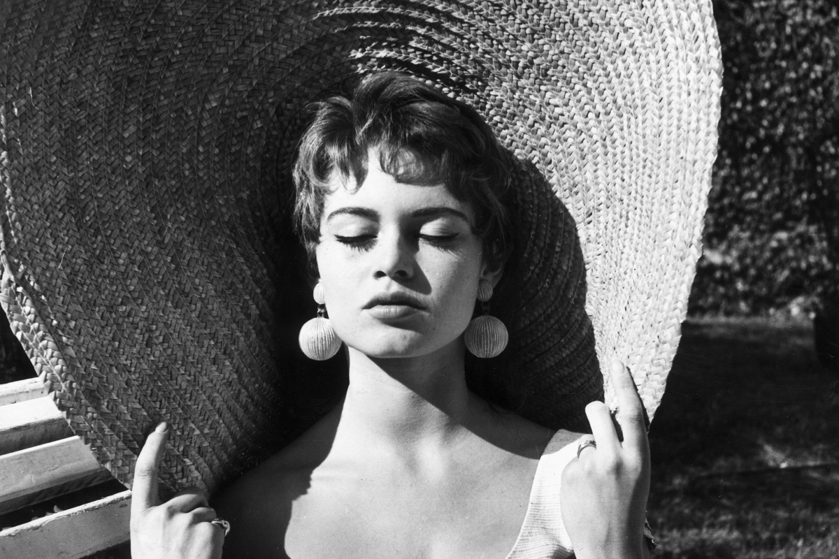 Brigitte Bardot chiude gli occhi e tiene il bordo del cappello di paglia mentre si crogiola al sole all'aperto. Bardot ha i capelli castani e indossa orecchini a goccia.