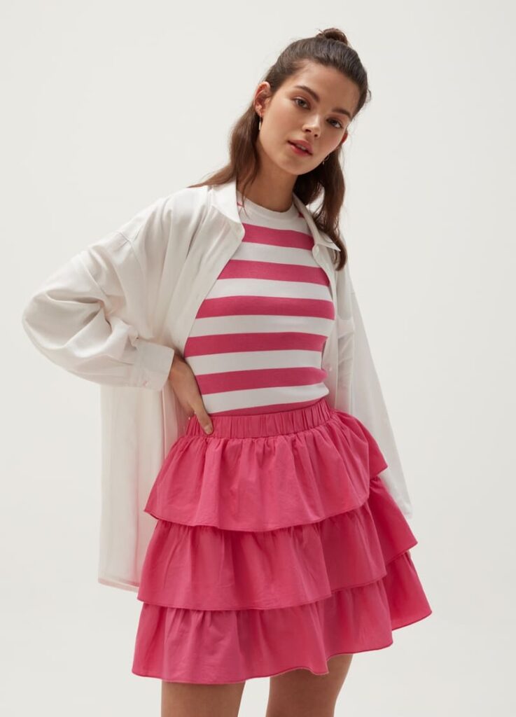 modella con maglia a righe rosa e bianca, camicia bianca aperta e gonna fucsia a balze su sfondo bianco