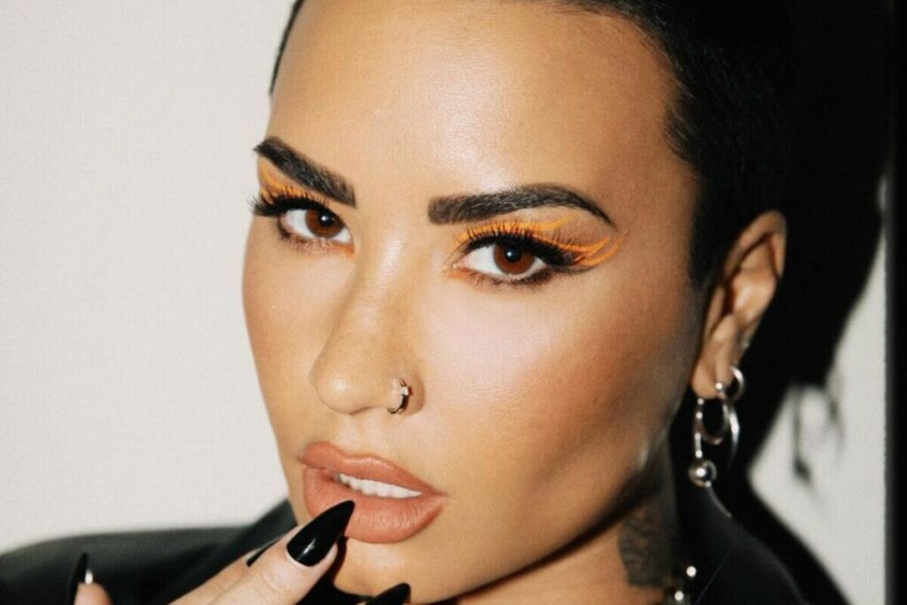 primo piano di Demi Lovato con capelli rasati, trucco grafico arancione ed unghie nere laccate