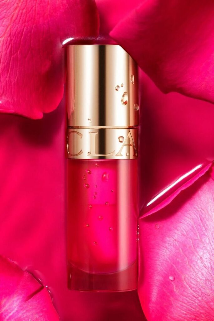 Lip Comfort Oil Clarins, olio labbra profumato su sfondo petali fiore