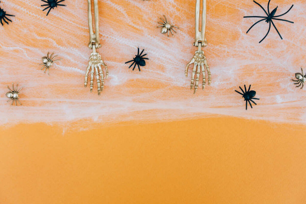 invito per halloween con sfondo di colore arancione, ragnatela bianca con ossa di mani e ragni appesi