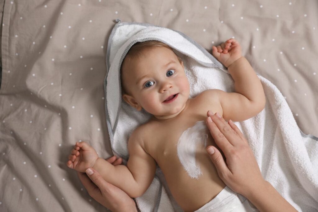 Bambino di pochi mesi con accappatoio, sdraiato mentre la madre gli spalma la crema sul corpo