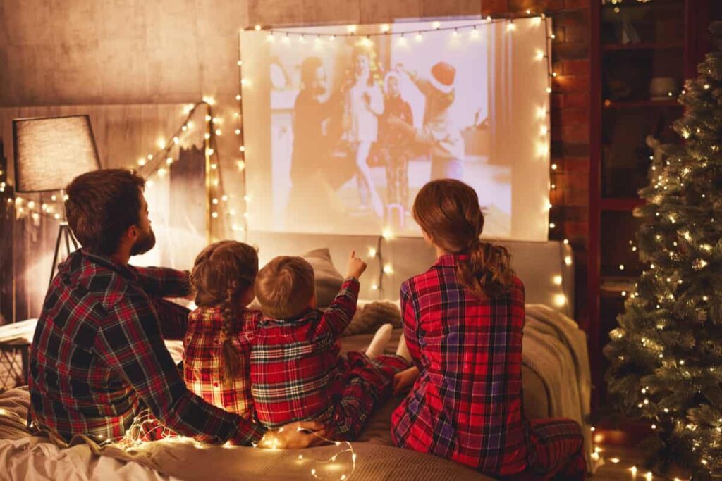 famiglia composta da mamma, papà, figlio e figlia con pigiami a quadri di colore rosso, davanti ad uno schermo a guardare film natalizi, con un albero di natale