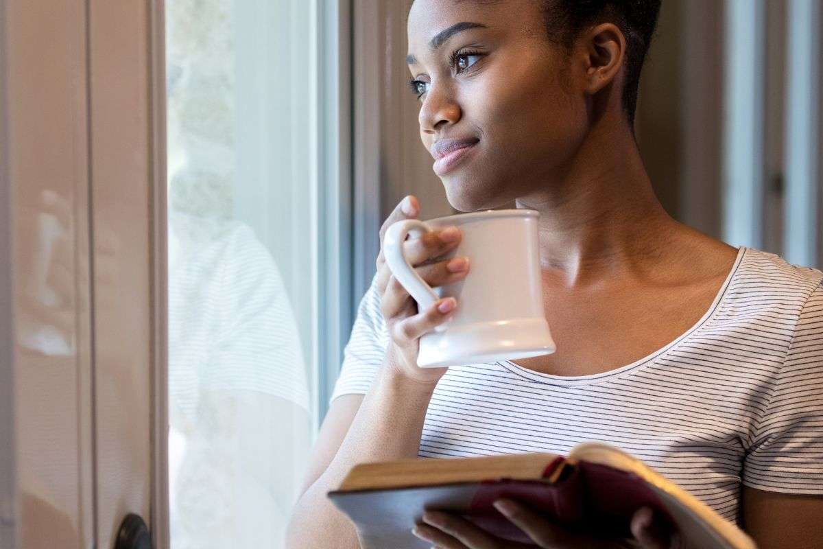persona con un libro e una tazza che guarda fuori dalla finestra